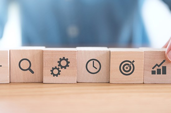 Stock photo of a hand placing a series of scrabble pieces in a line that include symbols of a light bulb, a target, and growth instead of letters.